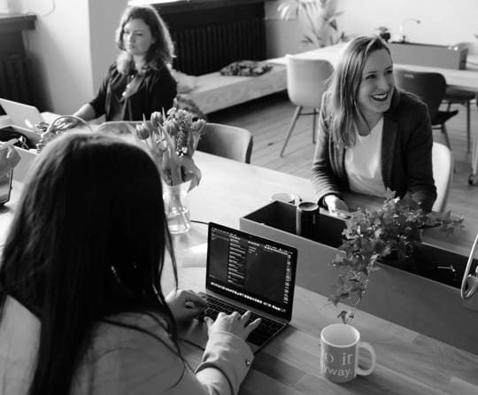 A couple of women sit in the cozy office. They are working with their laptops, one is laughing.